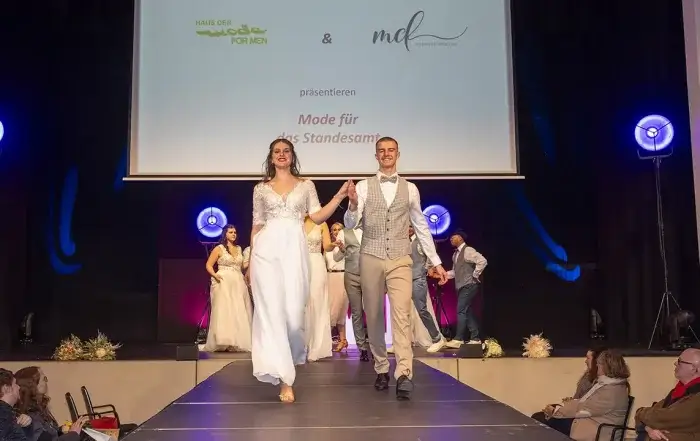 Eine Frau in einem weißen Kleid und ein Mann in einem grauen Anzug laufen lächelnd Hand in Hand über einen Laufsteg. Andere Models stehen im Hintergrund, während ein Hochzeits-DJ aus Thüringen Musik auflegt und eine Präsentationsleinwand über der Bühne leuchtet.