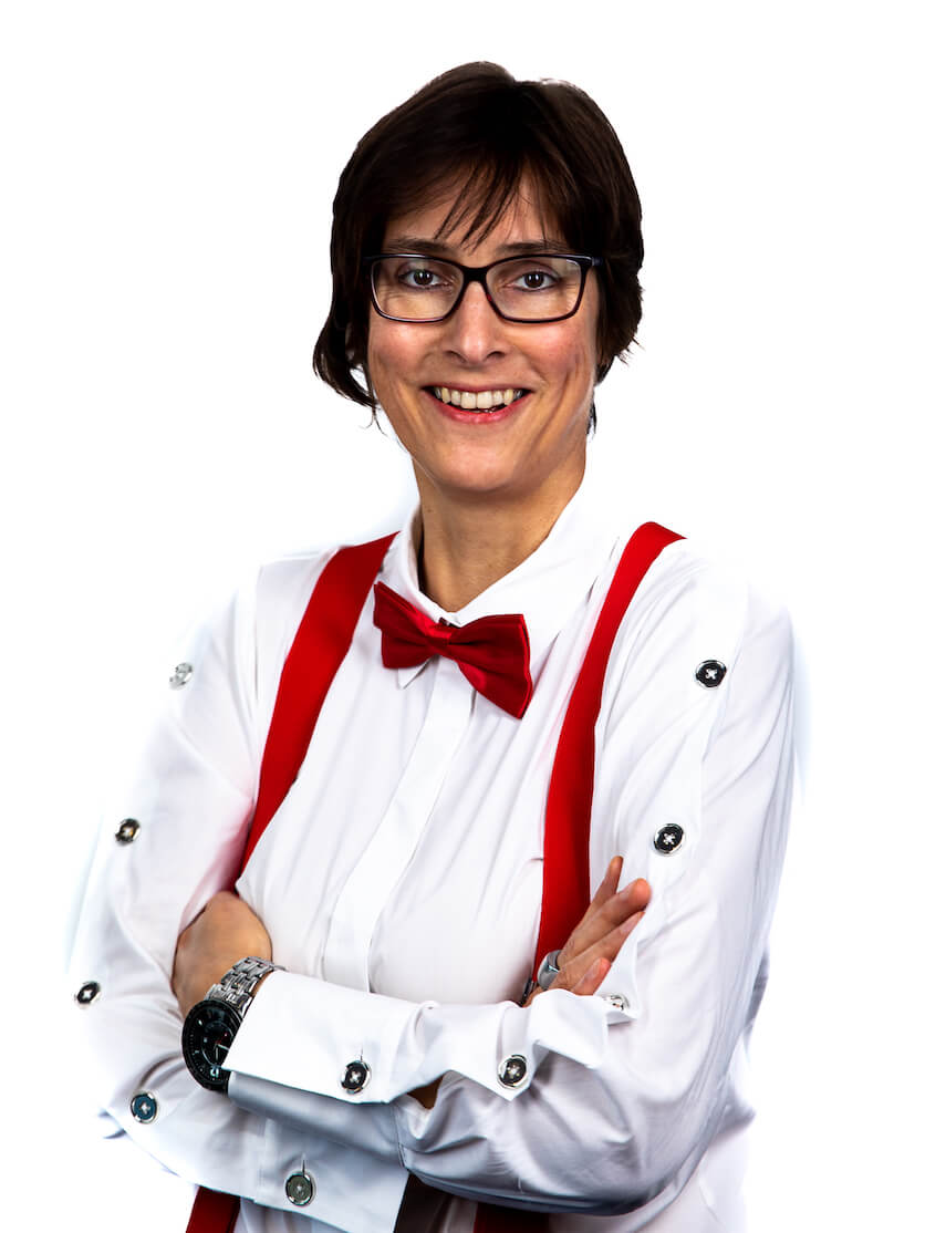 Eine Person mit kurzen dunklen Haaren und Brille lächelt mit verschränkten Armen, trägt ein weißes Hemd, rote Hosenträger und eine passende rote Fliege - perfekt gestylt für einen DJ für Hochzeit in Franken - vor einem schlichten weißen Hintergrund.