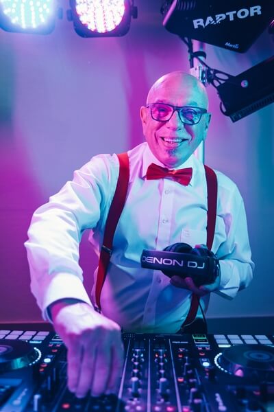 Ein älterer Mann mit Brille und roter Fliege lächelt, während er als Hochzeits-DJ in Thüringen auflegt. Er hält einen Denon DJ-Kopfhörer in der Hand und stellt die Regler an einem DJ-Mixer unter bunten Bühnenlichtern ein.