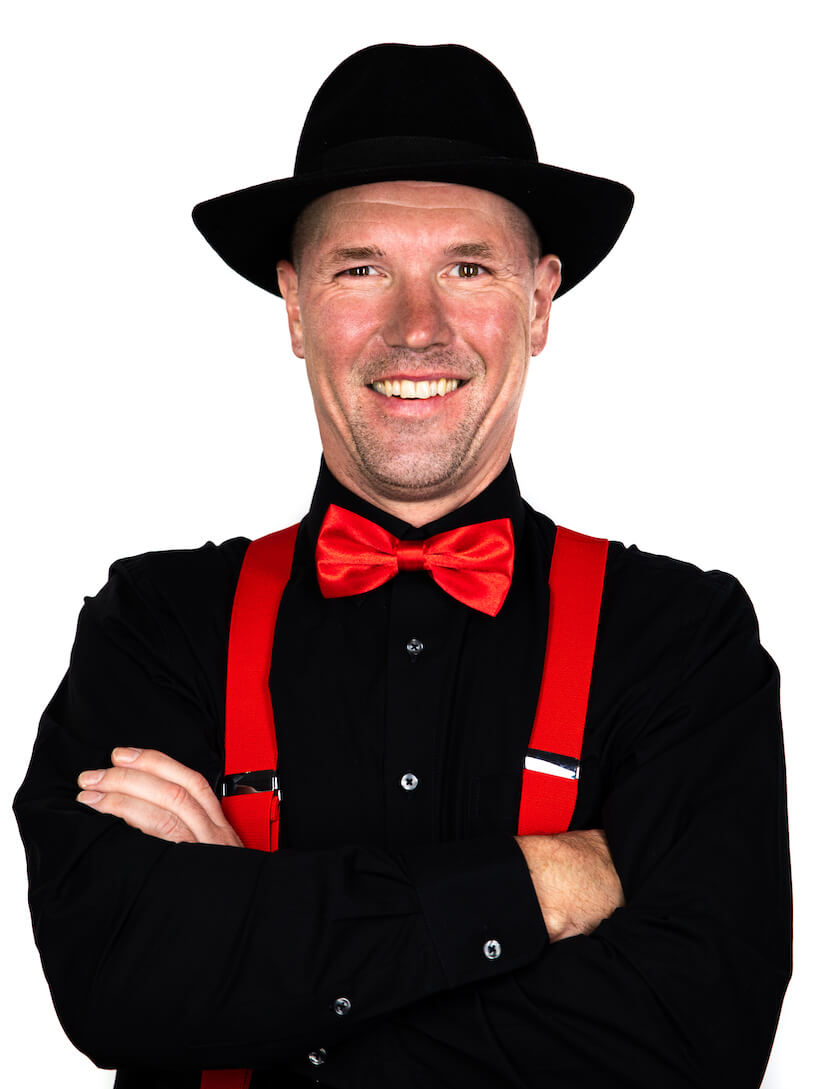Ein lächelnder Mann mit schwarzem Hemd, schwarzem Hut, roten Hosenträgern und roter Fliege steht mit verschränkten Armen vor einem weißen Hintergrund - HochzeitsDj Christian für Franken und Region Bamberg.