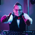 Ein lächelnder DJ mit Brille und Kopfhörern zeigt zwei Daumen nach oben hinter einem DJ-Mischpult, während im Hintergrund bunte Lichter leuchten - das ist HochzeitsDJ Ecky, der Energie und Spaß in jede Feier bringt.