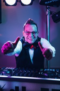 Hochzeits DJ in Franken und Thüringen ❤️ Die DJs mit der Roten Fliege ❤️ Ein lächelnder DJ mit Brille und Kopfhörern zeigt zwei Daumen nach oben hinter einem DJ-Mischpult, während im Hintergrund bunte Lichter leuchten - das ist HochzeitsDJ Ecky, der Energie und Spaß in jede Feier bringt.