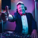 Ein gut gelaunter DJ für Hochzeit im Smoking und mit Kopfhörern gibt hinter einem Mischpult die Daumen hoch, im Hintergrund leuchten bunte Bühnenlichter - perfekt für Ihre Feier in Thüringen oder Franken.