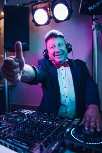 Hochzeits DJ in Franken und Thüringen ❤️ Die DJs mit der Roten Fliege ❤️ Ein gut gelaunter DJ für Hochzeit im Smoking und mit Kopfhörern gibt hinter einem Mischpult die Daumen hoch, im Hintergrund leuchten bunte Bühnenlichter - perfekt für Ihre Feier in Thüringen oder Franken.