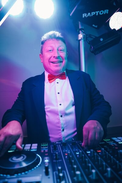 Ein lächelnder HochzeitsDJ im Smoking mit roter Fliege steht hinter einem DJ-Controller, beleuchtet von bunten Bühnenlichtern, bereit, auf Ihrer Veranstaltung in Thüringen Musik zu spielen.