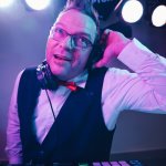 HochzeitsDJ Ecky aus Saalfeld Thüringen mit Kopfhörern, Weste und roter Fliege steht hinter einem DJ-Mischpult, lächelt und hält sich im hellen Bühnenlicht einen Kopfhörer ans Ohr.