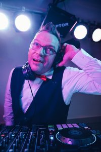Hochzeits DJ in Franken und Thüringen ❤️ Die DJs mit der Roten Fliege ❤️ HochzeitsDJ Ecky aus Saalfeld Thüringen mit Kopfhörern, Weste und roter Fliege steht hinter einem DJ-Mischpult, lächelt und hält sich im hellen Bühnenlicht einen Kopfhörer ans Ohr.