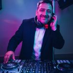 HochzeitsDJ Marco für die Party in Franken und Thüringen steht lächelnd im Anzug und roter Fliege hinter dem DJ-Mixer, hält Kopfhörer ans Ohr und genießt die Stimmung von der Bühne.