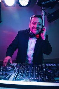 Hochzeits DJ in Franken und Thüringen ❤️ Die DJs mit der Roten Fliege ❤️ HochzeitsDJ Marco für die Party in Franken und Thüringen steht lächelnd im Anzug und roter Fliege hinter dem DJ-Mixer, hält Kopfhörer ans Ohr und genießt die Stimmung von der Bühne.
