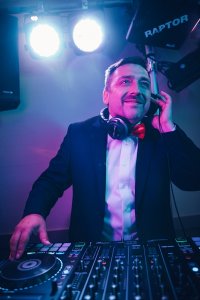 Hochzeits DJ in Franken und Thüringen ❤️ Die DJs mit der Roten Fliege ❤️ HochzeitsDJ Marco in Franken und Thüringen steht an einem DJ-Pult, lächelt in seinem Anzug mit Kopfhörern um den Hals und richtet ein Ohr unter bunten Bühnenlichtern auf den Beat aus.