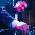 Ein DJ im Anzug und mit Kopfhörern lächelt, während er die Regler am Mischpult bedient, beleuchtet von bunten Bühnenlichtern - HochzeitsDJ Marco in Thüringen und Franken bringt unvergessliche Musik auf Ihre Feier.