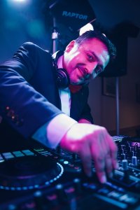 Hochzeits DJ in Franken und Thüringen ❤️ Die DJs mit der Roten Fliege ❤️ Ein DJ im Anzug und mit Kopfhörern lächelt, während er die Regler am Mischpult bedient, beleuchtet von bunten Bühnenlichtern - HochzeitsDJ Marco in Thüringen und Franken bringt unvergessliche Musik auf Ihre Feier.
