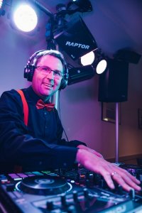 Hochzeits DJ in Franken und Thüringen ❤️ Die DJs mit der Roten Fliege ❤️ Ein lächelnder Hochzeits-DJ mit Brille, Kopfhörern, schwarzem Hemd und roter Fliege steht an einem Mischpult mit bunten Lichtern und Lautsprechern im Hintergrund und ist bereit, Energie in jede Thüringer Feier zu bringen.