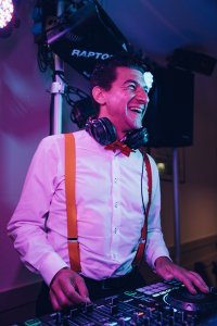 Hochzeits DJ in Franken und Thüringen ❤️ Die DJs mit der Roten Fliege ❤️ Ein lächelnder Hochzeits-DJ mit weißem Hemd, roten Hosenträgern und Kopfhörern um den Hals steht an einem DJ-Mischpult, das mit bunten Lichtern beleuchtet ist - perfekt für Hochzeiten in Thüringen oder Franken.