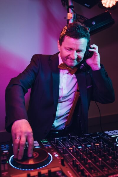 Ein DJ in Anzug und Fliege mischt Musik an einem DJ-Controller, hält Kopfhörer an ein Ohr unter bunter Bühnenbeleuchtung - perfekt als Ihr Hochzeit DJ in Thüringen.
