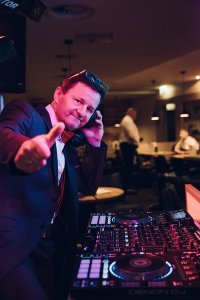 Hochzeits DJ in Franken und Thüringen ❤️ Die DJs mit der Roten Fliege ❤️ Ein DJ für Hochzeit in Thüringen, stilvoll im Anzug und mit Sonnenbrille, steht hinter einem DJ-Mischpult, hält Kopfhörer ans Ohr und zeigt in die Kamera in einer lebhaften, schwach beleuchteten Innenumgebung.