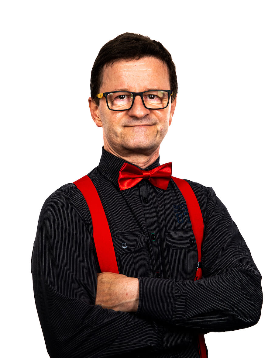 Ein Mann mit kurzen dunklen Haaren und Brille, schwarzem Hemd, roten Hosenträgern und roter Fliege steht mit verschränkten Armen und leichtem Lächeln - Ihr stilvoller HochzeitsDJ für Franken und Thüringen - vor einem schlichten weißen Hintergrund.