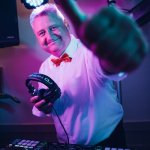 Ein lächelnder Hochzeits-DJ in weißem Hemd und roter Fliege hält Kopfhörer in der Hand und gestikuliert in die Kamera. Er steht hinter einem DJ-Mischpult unter bunten Lichtern.