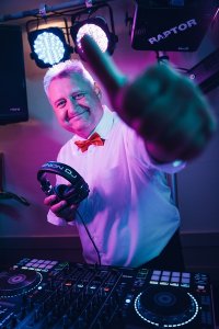 Hochzeits DJ in Franken und Thüringen ❤️ Die DJs mit der Roten Fliege ❤️ Ein lächelnder Hochzeits-DJ in weißem Hemd und roter Fliege hält Kopfhörer in der Hand und gestikuliert in die Kamera. Er steht hinter einem DJ-Mischpult unter bunten Lichtern.