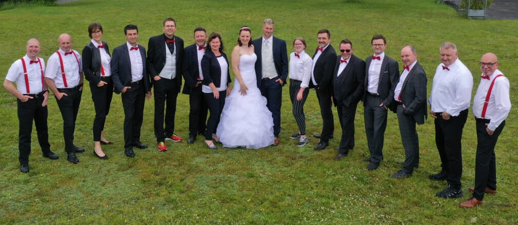 Braut und Bräutigam stehen in der Mitte einer formell gekleideten Gruppe, einige mit Roten Fliege und roten Hosenträgern. Das sind die HochzeitsDJs mit der Roten Fliege. Alle lächeln, schauen in die Kamera und posieren im Freien auf der grünen Wiese.