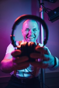Hochzeits DJ in Franken und Thüringen ❤️ Die DJs mit der Roten Fliege ❤️ Ein lächelnder Mann im weißen Hemd hält Kopfhörer in die Kamera, die in kräftiges lila und blaues Licht getaucht sind - und fängt die lebendige Energie von HochzeitsDj Christian für Franken und in Bamberg ein.