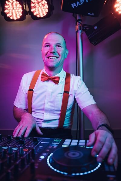 Ein lächelnder Hochzeits-DJ aus Franken, der ein weißes Hemd, rote Hosenträger und eine rote Fliege trägt, tritt an einem Mischpult unter buntem Bühnenlicht auf.