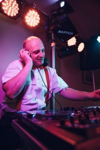 Hochzeits DJ in Franken und Thüringen ❤️ Die DJs mit der Roten Fliege ❤️ Ein Hochzeits-DJ mittleren Alters in weißem Hemd und roten Hosenträgern tritt in Franken auf, trägt Kopfhörer und bedient ein Mischpult unter buntem Bühnenlicht.