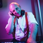 Ein lächelnder Mann mit weißem Hemd, roten Hosenträgern und Kopfhörern steht unter bunten Bühnenlichtern an einem DJ-Mischpult und scheint es zu genießen, als Hochzeits-DJ in Franken Musik aufzulegen.