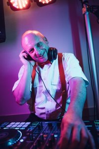 Hochzeits DJ in Franken und Thüringen ❤️ Die DJs mit der Roten Fliege ❤️ Ein lächelnder Mann mit weißem Hemd, roten Hosenträgern und Kopfhörern steht unter bunten Bühnenlichtern an einem DJ-Mischpult und scheint es zu genießen, als Hochzeits-DJ in Franken Musik aufzulegen.