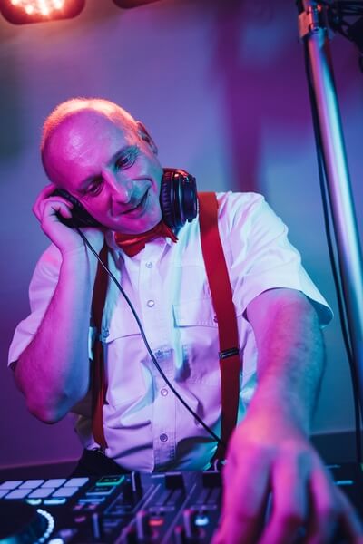Ein lächelnder älterer Mann, der perfekte Hochzeits-DJ, trägt Kopfhörer, ein weißes Hemd, rote Hosenträger und eine Fliege, während er in leuchtendem Lila und Blau auflegt und die Regler eines DJ-Mixers bedient - ideal für jede Thüringer Feier.