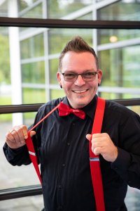 Hochzeits DJ in Franken und Thüringen ❤️ Die DJs mit der Roten Fliege ❤️ Ein lächelnder Mann mit Brille und kurzer Irokesenfrisur, gekleidet als Hochzeits-DJ mit schwarzem Hemd, roter Fliege und roten Hosenträgern, steht in einer Halle in Thüringen und hält seine Hosenträger mit beiden Händen vor großen Fenstern.