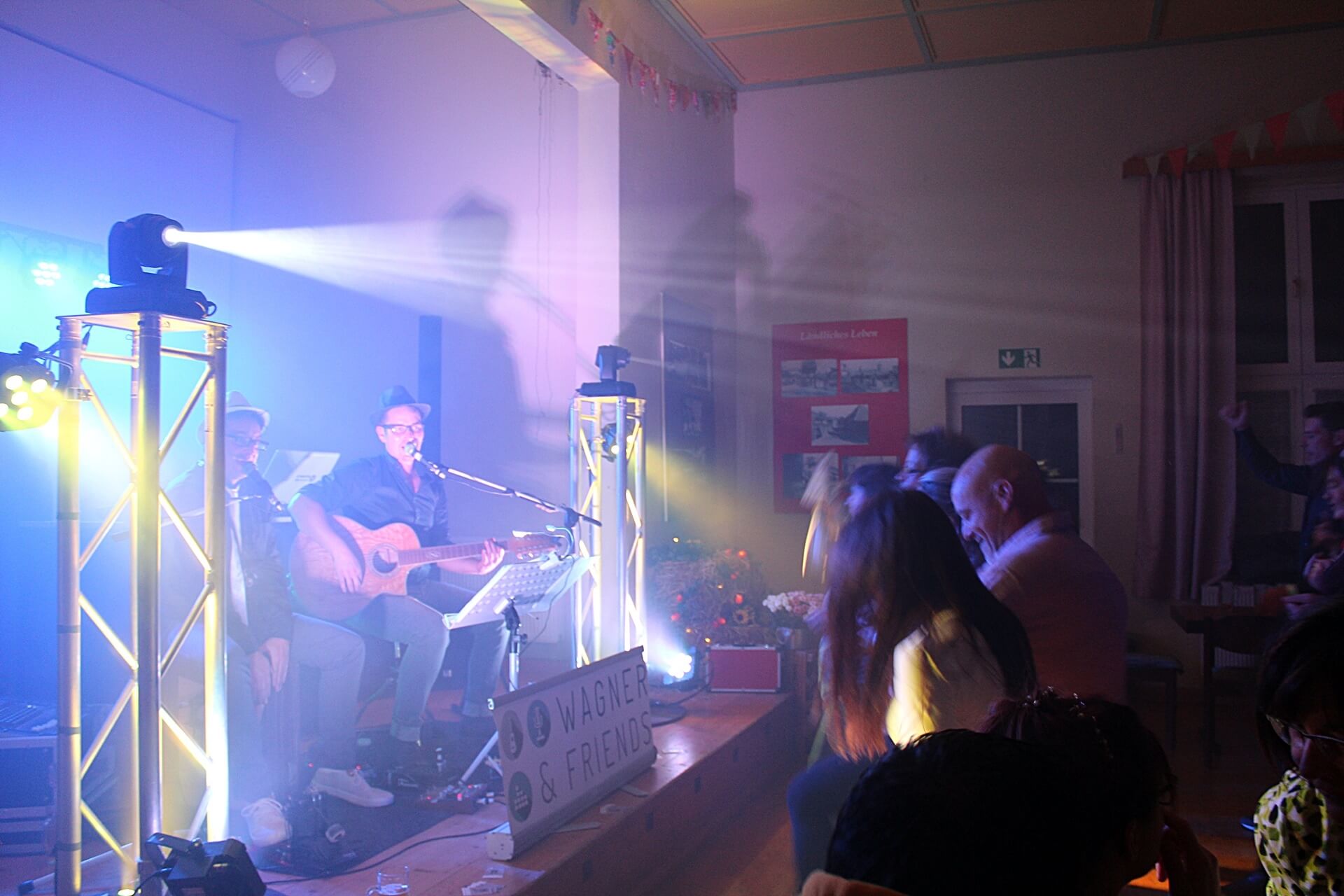 Hochzeits DJ in Franken und Thüringen ❤️ Die DJs mit der Roten Fliege ❤️ Eine Band spielt live auf einer kleinen, hell erleuchteten Bühne mit Gitarren und Mikrofonen. Das Publikum genießt die Musik in einer gemütlichen Halle, während auf einem Schild WAGNER & FRIENDS steht - die perfekte Inspiration für alle, die einen Hochzeits-DJ suchen.