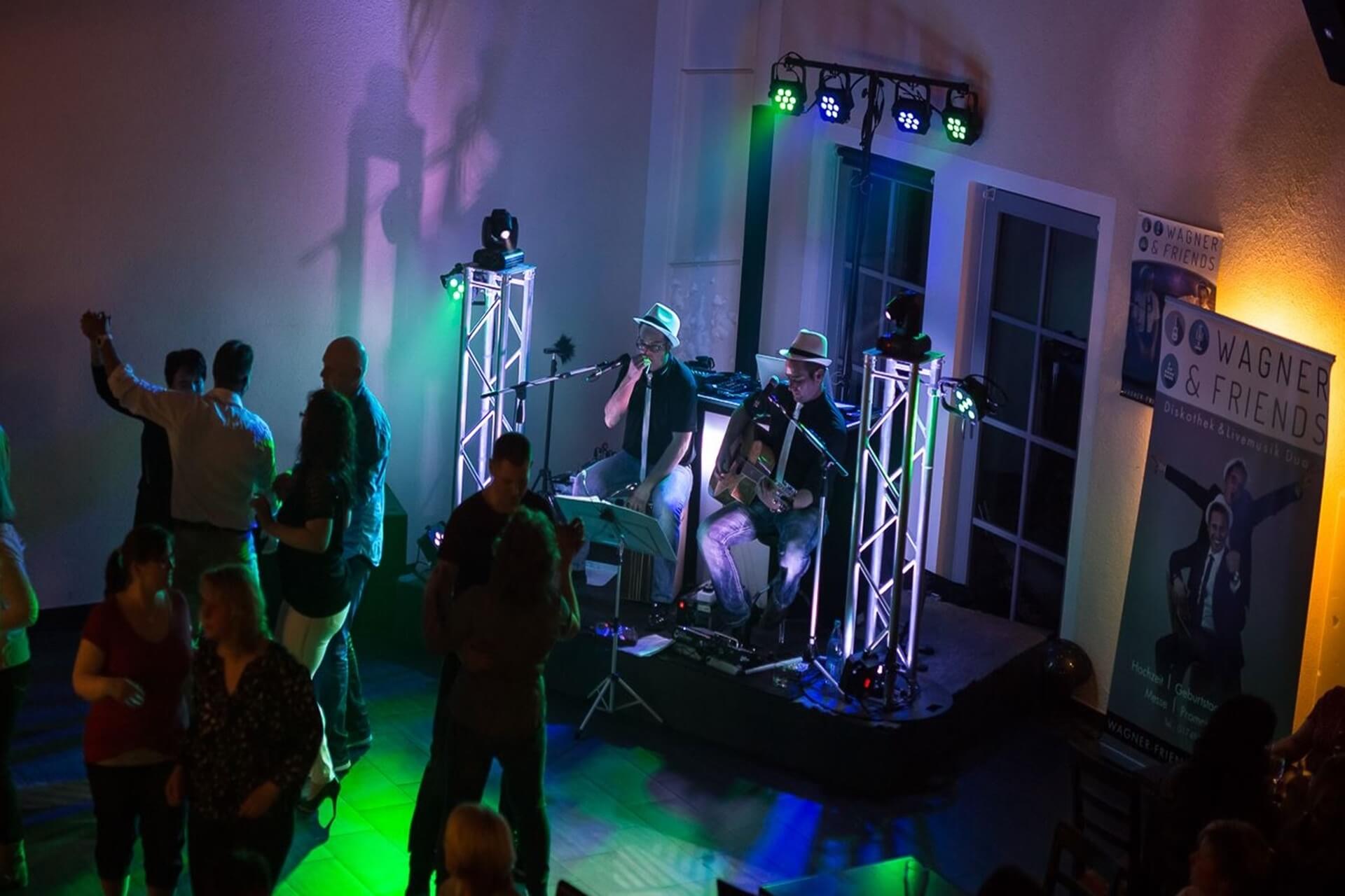 Hochzeits DJ in Franken und Thüringen ❤️ Die DJs mit der Roten Fliege ❤️ Eine kleine Band spielt auf der Bühne mit bunten Lichtern, während die Leute auf dem Boden tanzen und sich unterhalten. Auf dieser lebhaften Indoor-Veranstaltung in Franken ist ein Werbebanner für einen DJ für Hochzeit zu sehen, das die festliche Atmosphäre noch verstärkt.